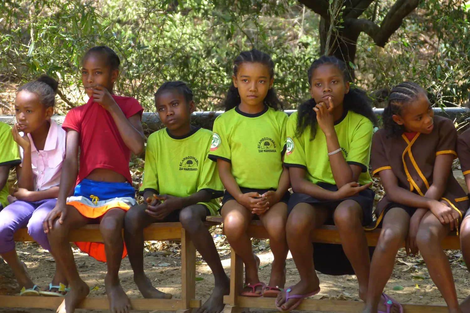 A group of schoolchildren sit on a wooden bench outdoors, some wearing green uniforms with the school name and logo.
