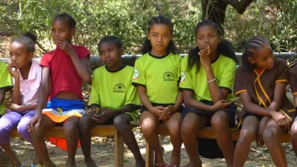 A group of schoolchildren sit on a wooden bench outdoors, some wearing green uniforms with the school name and logo.
