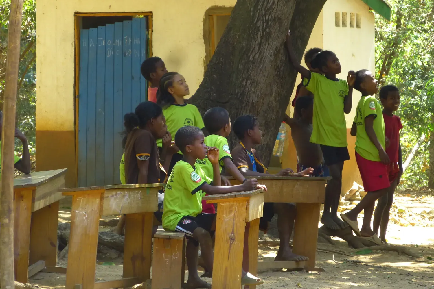 Children in green and brown school uniforms gather around desks and trees in an outdoor classroom setting.