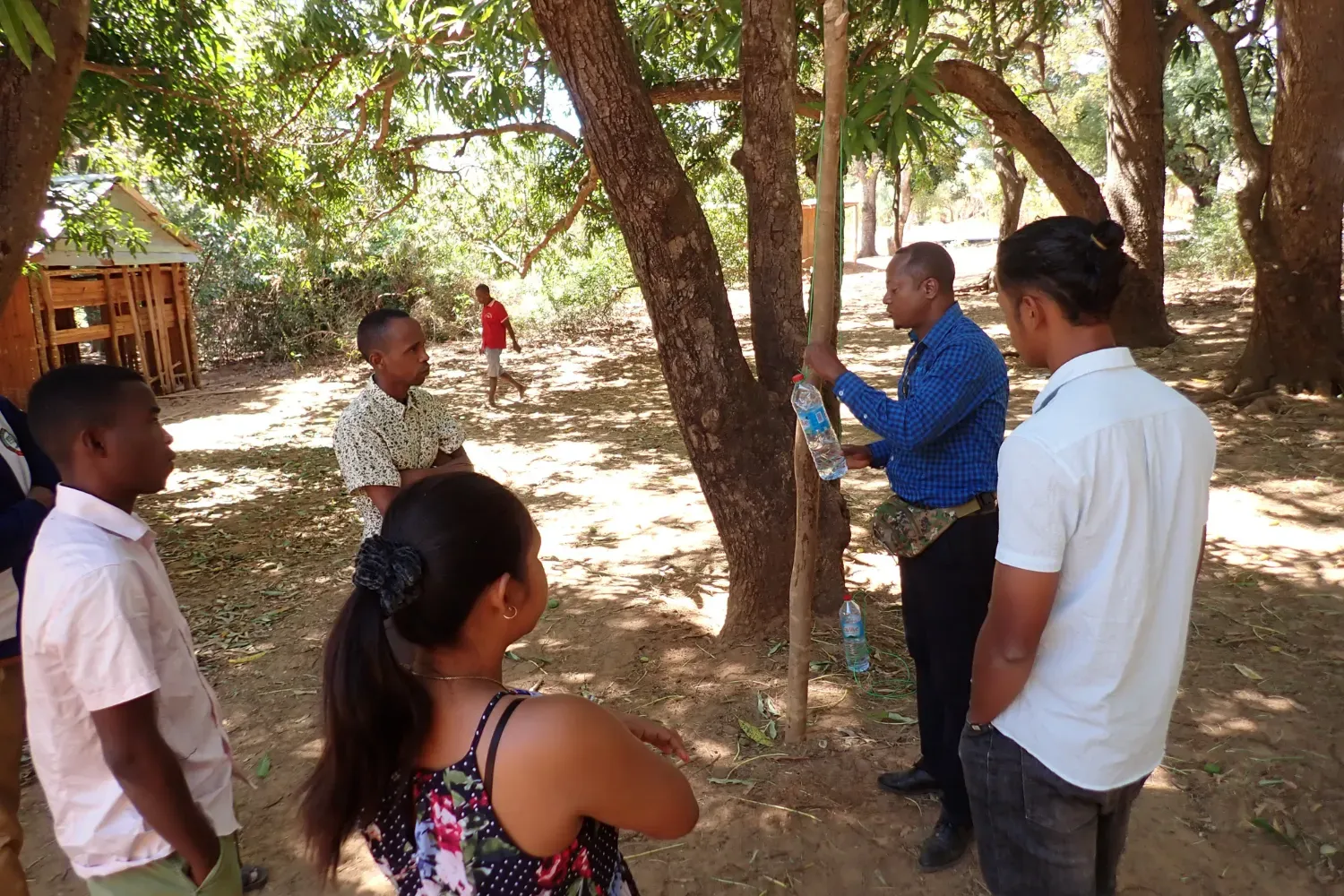 A group of adults stand in a shaded forest area, observing a demonstration involving a water bottle and bamboo stick.