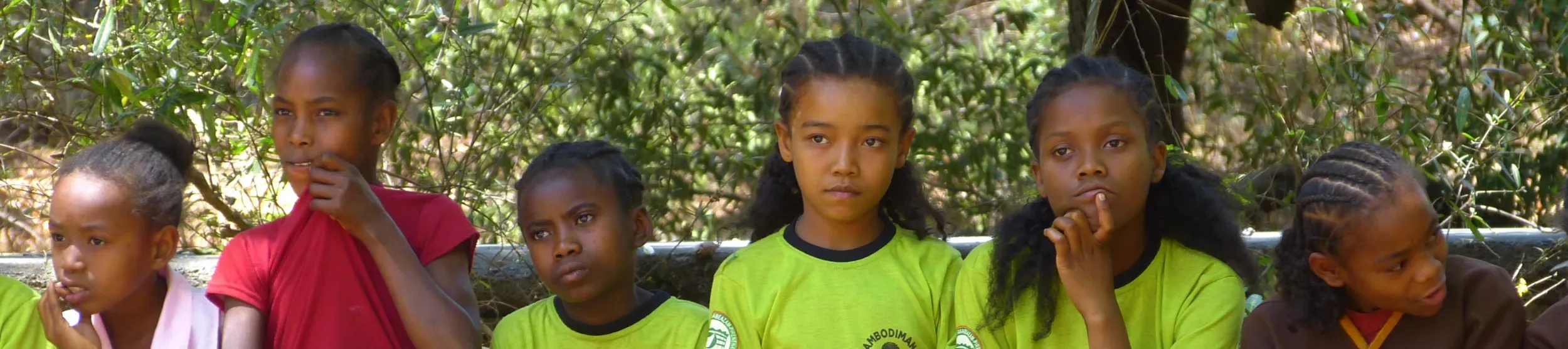 A group of schoolchildren sit on a wooden bench outdoors, some wearing green uniforms with the school name and logo.
