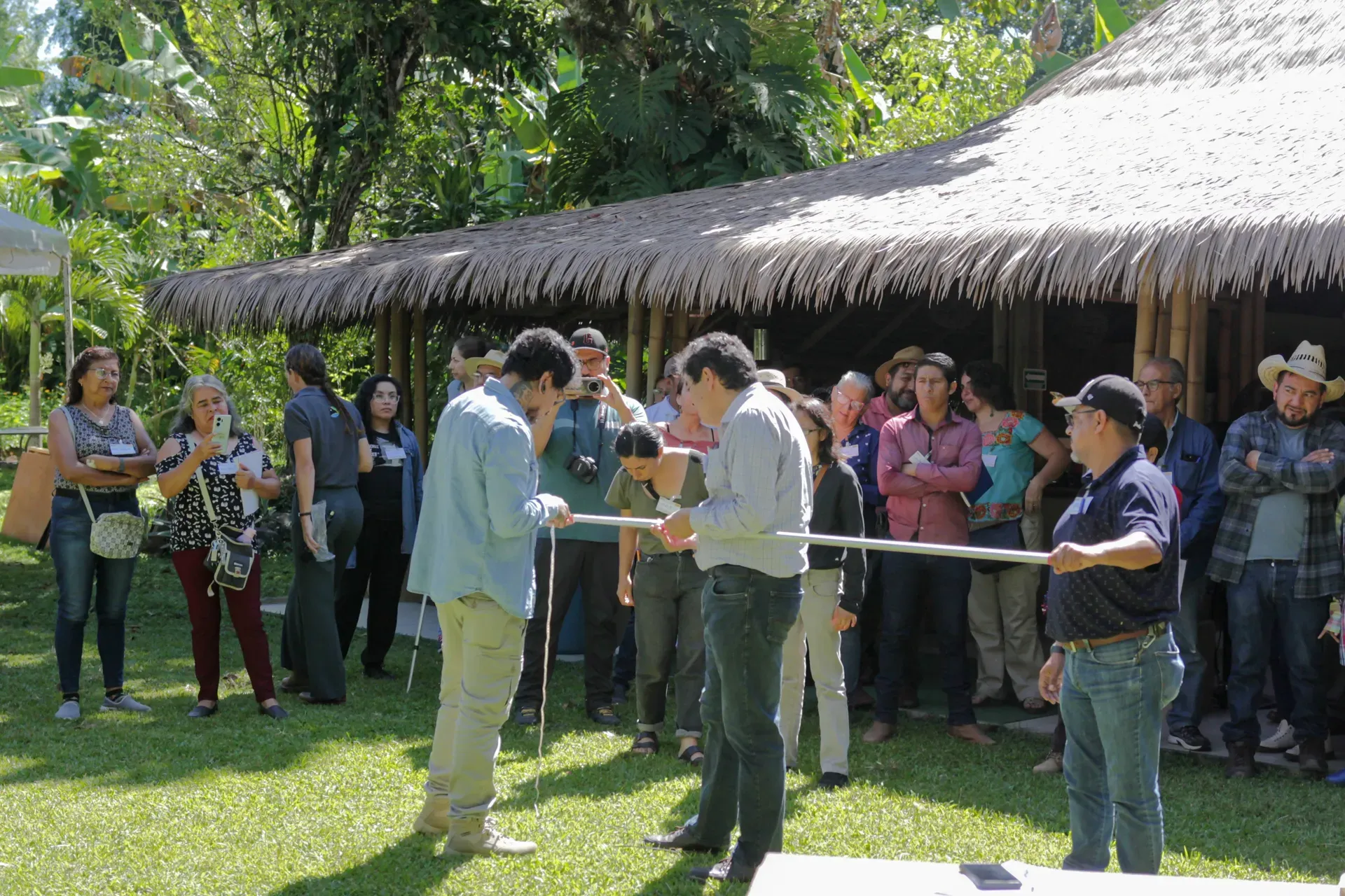 A small group of people are being trained to use some specialist kit while a larger group of onlookers watch from the shade cover of an open building