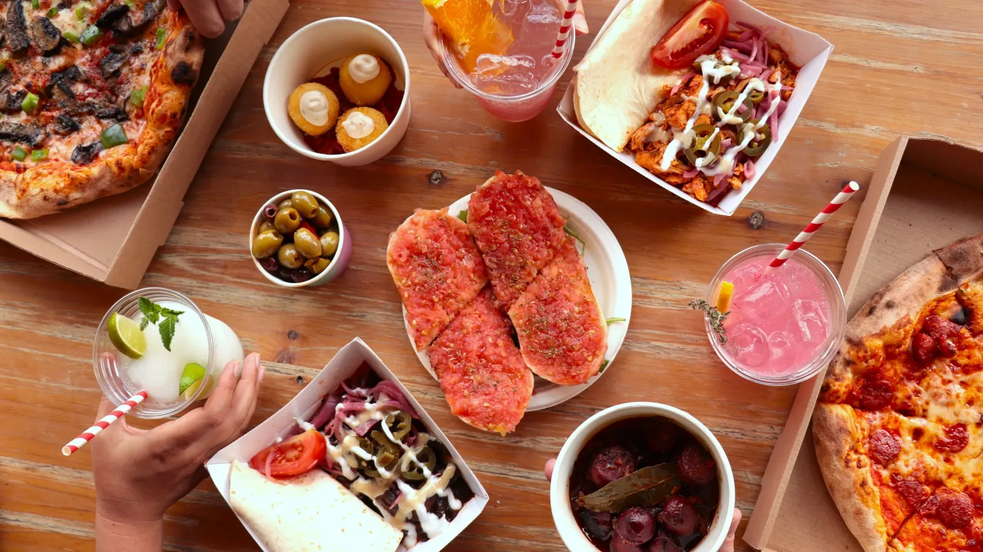 Top-down view of a table with pizza, shawarma wraps, bruschetta, olives, fried bites, sausages, and colourful drinks with straws.