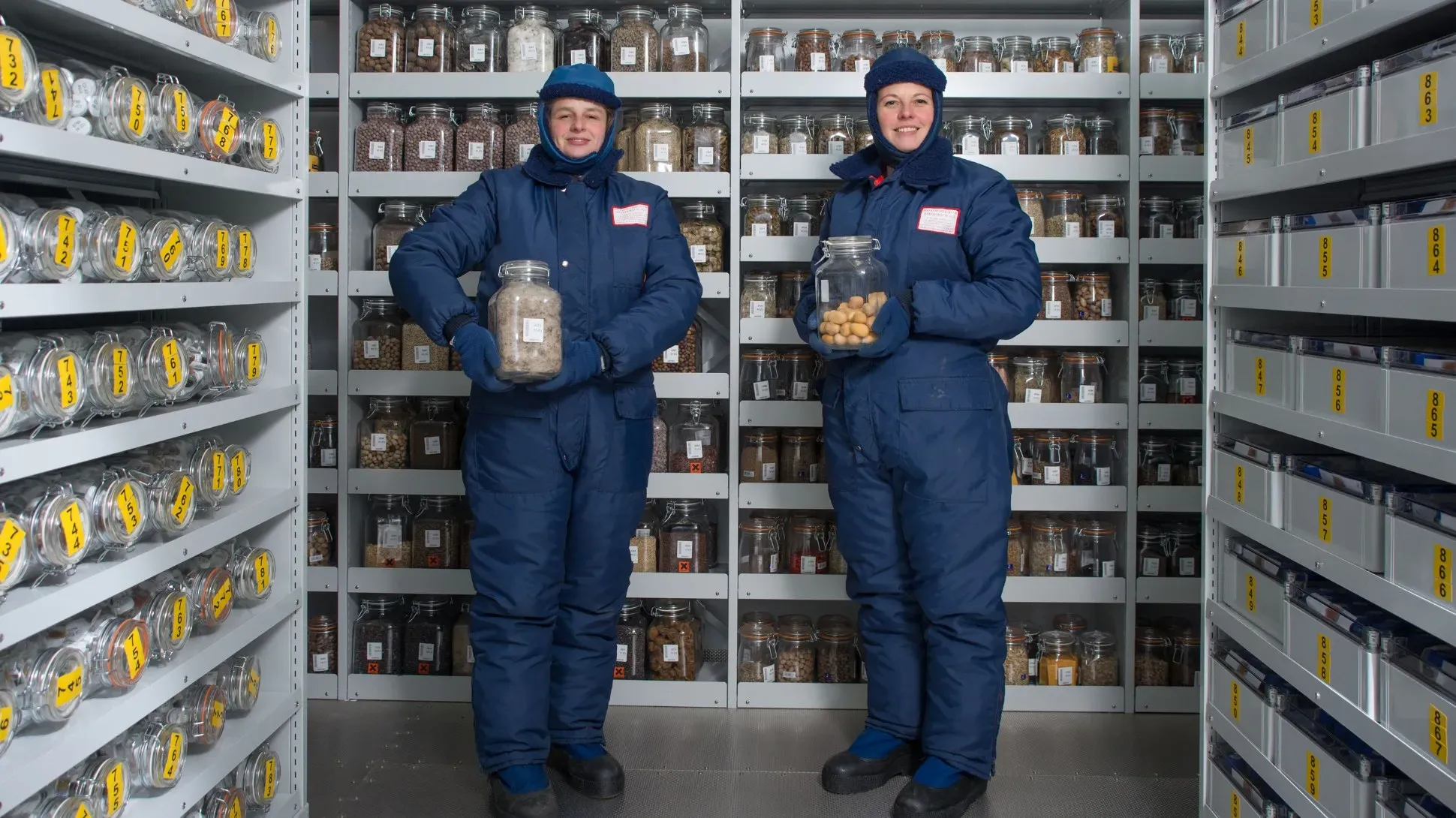 Two scientists in blue suits holding jars in the vault of the Millennium Seed Bank